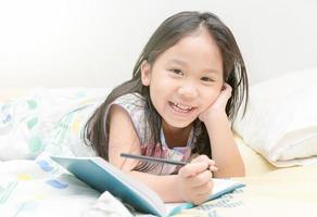 linda asiático niña sonrisa y escritura a diario en el cama foto