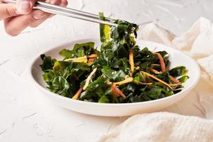 homemade seaweed salad or kelp in the white dish being pinched with chopsticks on white table background photo