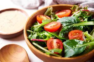 green salad from green leaves and cherry tomato in wooden bowl on white stone background photo