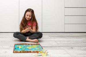 niña se sienta en el piso a hogar, sostiene jugando tarjetas en su manos, un tablero juego es puesto fuera en el piso foto