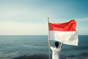 un hombre participación un rojo y blanco Indonesia bandera mirando a el océano. ai generado foto