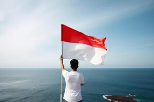 un hombre participación un rojo y blanco Indonesia bandera mirando a el océano. ai generado foto