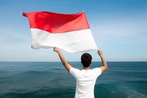 un hombre participación un rojo y blanco Indonesia bandera mirando a el océano. ai generado foto