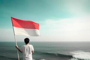 un hombre participación un rojo y blanco Indonesia bandera mirando a el océano. ai generado foto