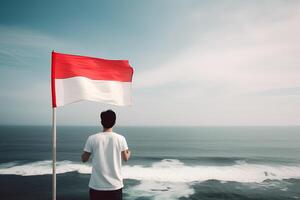 un hombre participación un rojo y blanco Indonesia bandera mirando a el océano. ai generado foto