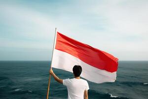 A man holding a red and white Indonesia flag looking at the ocean. photo