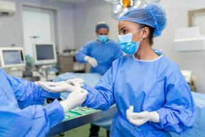 joven enfermera con máscara protectora y ropa de trabajo que ayuda al cirujano con guantes mientras ambos se preparan para la operación foto