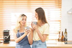 happy lesbian couple holding cups of coffee in kitchen. Couple of lesbian Girls Enjoy coffee at home taking about something. Two young adult beautiful women drink tea in modern kitchen. photo