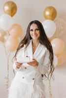 joven caucásico mujer participación un cumpleaños pastel con un vela en un elegante blanco vestir con plumas foto
