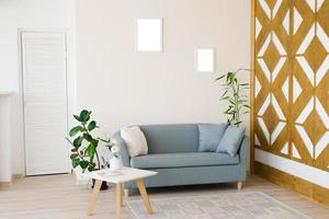 un polvoriento azul sofá con almohadas, un blanco café mesa, en conserva plantas en un brillante acogedor vivo habitación. Bosquejo marcos para carteles en el pared foto