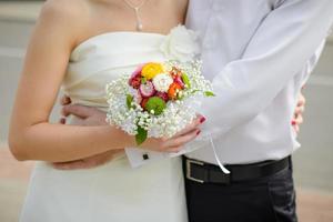 linda pequeño nupcial ramo de flores de el novia en el manos de el novia, el novia es abrazado por el novio foto