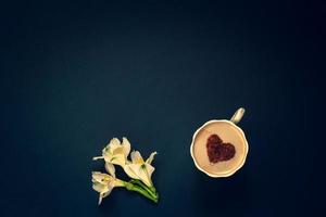 Minimalistic flat lay. Several buds of flowers and coffee with cream and cinnamon in the form of a heart in a mug on a dark blue background. White with yellow petals. Vignetting. Copy space on top. photo