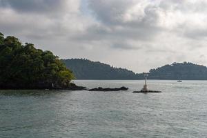 A small white lighthouse on the stones near the island on the river. A small boat is visible in the distance. On the slopes of the mountains houses are visible. Horizontal. photo