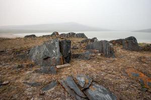 Beautiful black rocks sticking out of the ground with a lake and hills, foggy morning. Stones with red moss like rust. Dead grass. Grey sky. Copy space. photo