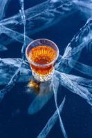 A glass of whiskey stands on beautiful ice with deep cracks. Orange liquid on a blue background. Top view from the side. Bizarre white deep cracks in the ice. Vertical. photo