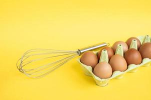 Tray with chicken eggs and a metal whisk on a yellow background. Prepare ingredients for cooking photo