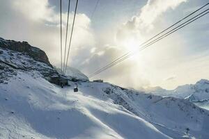 Gondola lift moving over snowcapped mountains under cloudy sky photo