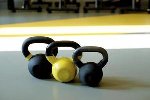 three kettlebells stand in a row on a gray floor in a bright fitness room. Yellow kettlebell, black kettlebell photo