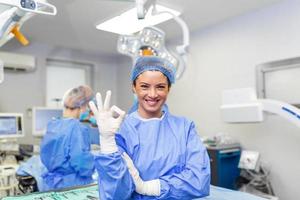 Portrait of female woman nurse surgeon OR staff member dressed in surgical scrubs gown mask and hair net in hospital operating room theater making eye contact smiling showing ok sign photo