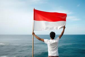 un hombre participación un rojo y blanco Indonesia bandera mirando a el océano. ai generado foto
