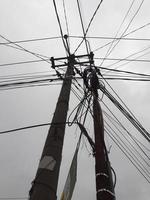 The messy power lines on power poles are photographed from below against a gray sky as a background. photo