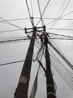 The messy power lines on power poles are photographed from below against a gray sky as a background. photo