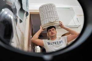 Man with basket on head, view from washing machine inside. Male does laundry daily routine. photo