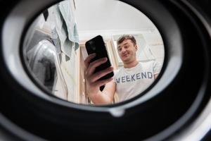 Man with mobile phone, view from washing machine inside. Male does laundry daily routine. photo