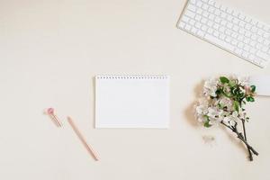 Open notebook with a blank white sheet of paper for copy space, keyboard, pen and white flowers on a beige background. Flat lay workspace photo