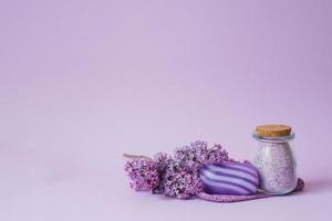 Cosmetic spa set for body care. Sea salt in glass jar, organic soap, washcloth glove and lilac flowers on purple background with copy space photo