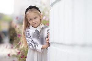 A beautiful little blonde girl stands by a brick wall in the summer. A child in town. photo