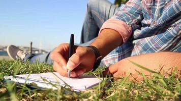 persoon aan het liegen Aan gras en nemen aantekeningen papier Aan klembord, Mens werken en nemen Notitie in een groen stad park Aan een zonnig dag, selectief focus video