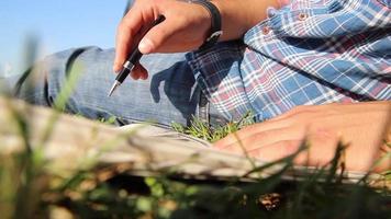 joven hombre leyendo periódico a jardín parque en un soleado día, hombre acostado en césped en ciudad parque leyendo periódico y participación Nota con bolígrafo, selectivo atención video