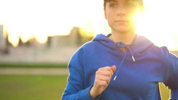 femme avec sans fil écouteurs et téléphone intelligent choisit la musique et court par le stade à le coucher du soleil. lent mouvement video