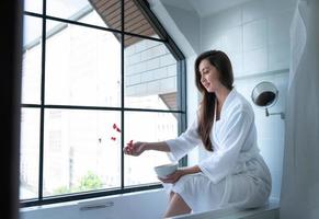 Woman sitting on bathtub wearing white bathrobe in modern bathroom at home with rose leaves to enhance the fragrance of the bath atmosphere photo