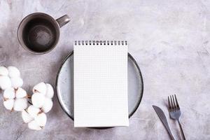 Mockup of a notebook on a plate, an empty cup and a branch of cotton on a gray background top view photo