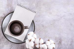 Empty ceramic mug and notebook on a plate and a branch of cotton on a gray background top view photo