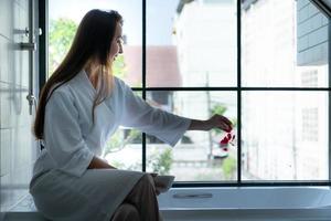 Woman sitting on bathtub wearing white bathrobe in modern bathroom at home with rose leaves to enhance the fragrance of the bath atmosphere photo