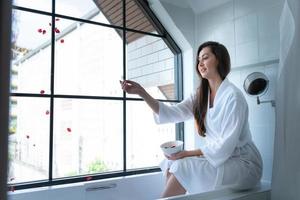 Woman sitting on bathtub wearing white bathrobe in modern bathroom at home with rose leaves to enhance the fragrance of the bath atmosphere photo