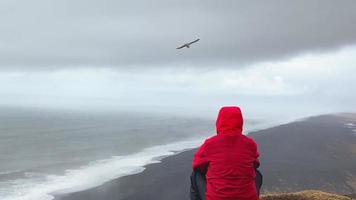 Mann im rot Jacke sitzen nachdenklich aussehen beim atlantisch Ozean Wellen. berühmt ikonisch Cliff Standpunkt Über reynisfjara schwarz Sand Strand. Person sieht aus zum Richtung und Zweck auf Reisen video