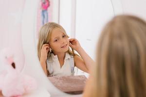 Happy child girl of seven years old puts on earrings in front of the mirror photo