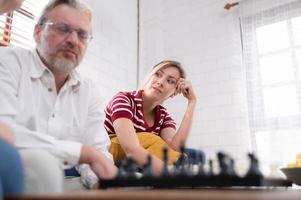 In the living room of the house, an elderly couple sits and relaxes. to begin playing chess together with a chess board with a daughter cheering beside him photo