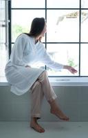 Woman sitting on bathtub wearing white bathrobe in modern bathroom at home with rose leaves to enhance the fragrance of the bath atmosphere photo