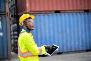 worker man in protective safety jumpsuit uniform with yellow hardhat and use laptop check container at cargo shipping warehouse. transportation import,export logistic industrial service photo