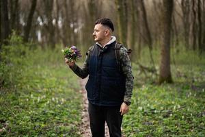 Portrait of man with backpack holding bouquet of spring flowers on forest trail. Love to nature. photo