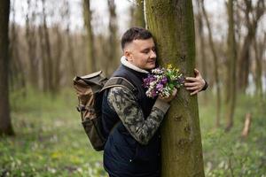 Portrait of man with backpack holding bouquet of spring flowers and hug tree on forest. Love to nature. photo