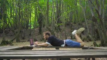 Young adventurer working with laptop in nature. Young man lying on a bench in the forest works with a laptop. video