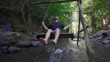 giovane uomo oscillante su un' swing sopra il acqua. lento movimento. acqua tocchi il piedi di il giovane uomo oscillante su il oscillazione. video