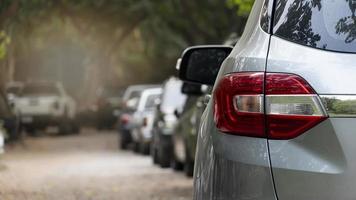 Rear view of a car in a residential parking lot. Visible taillights of a gray car and blurred of mirror wing. Long row of cars parked in quiet neighborhood. Blurred of shade of tall trees beside road. photo