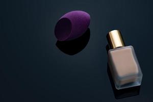 Purple makeup sponge on dark background. Soft makeup cosmetic sponge. Flat-ended makeup sponge with blur makeup foundation bottle. Liquid beige make-up foundation and beauty blender. Cosmetic product. photo